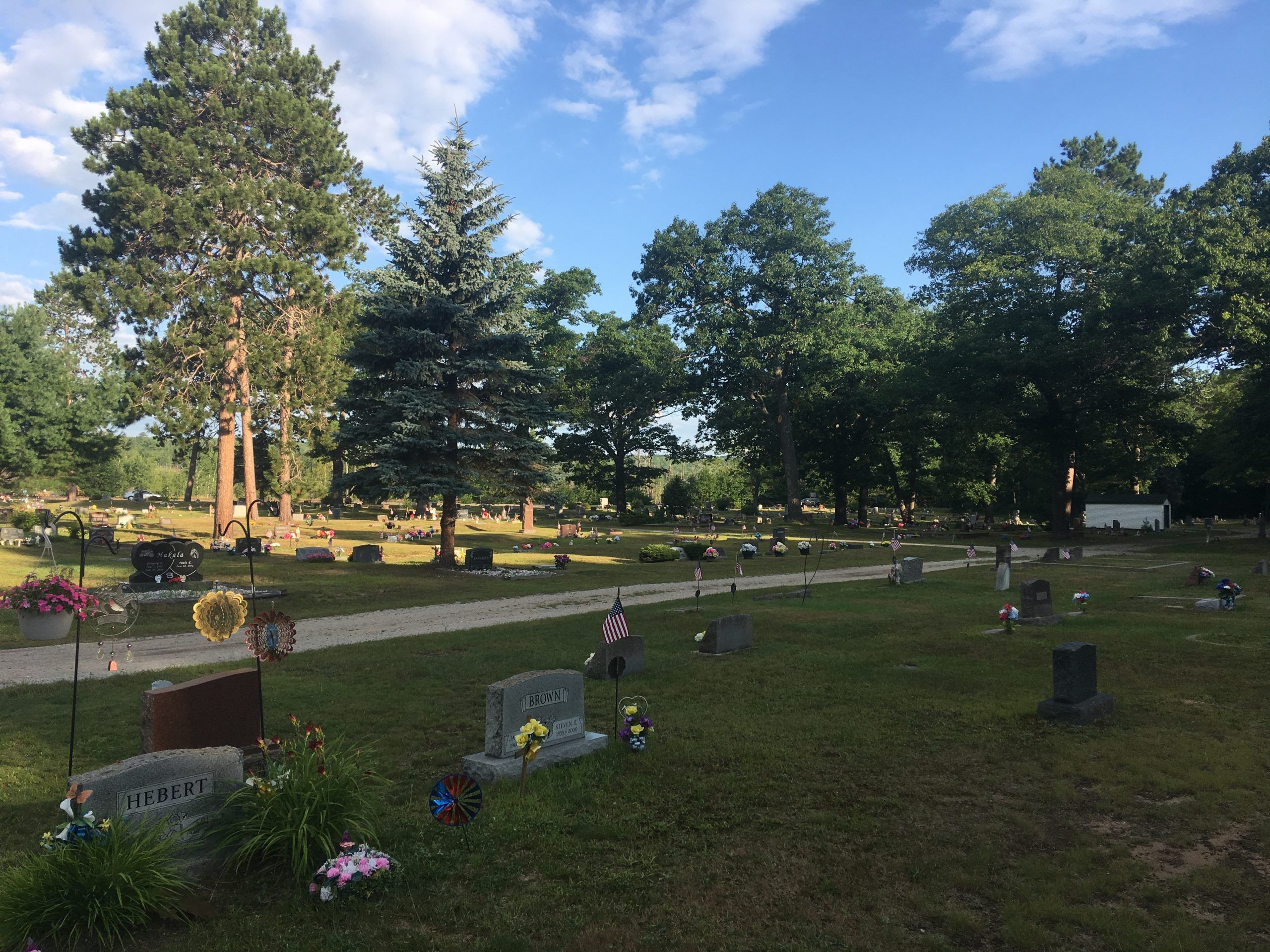 View of cemetery driving path and headstones with flowers and decorations. Trees are also throughout the cemetery.